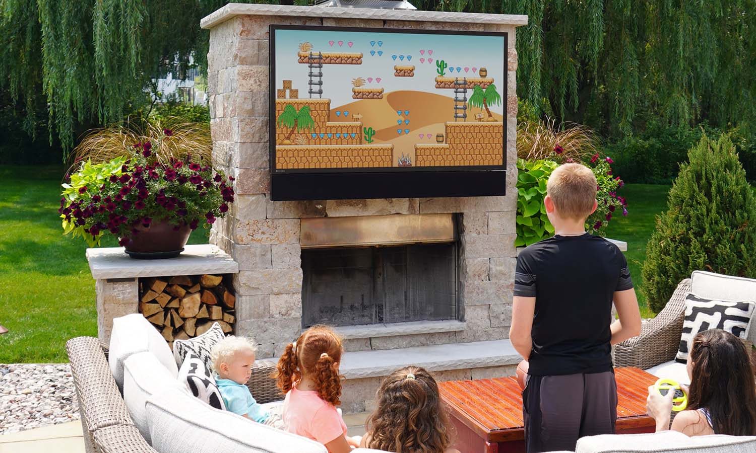 A father and daughter sitting on a cozy outdoor couch at night, enjoying quality time near a fireplace with string lights above, while a TV on the stone wall displays a colorful video game.